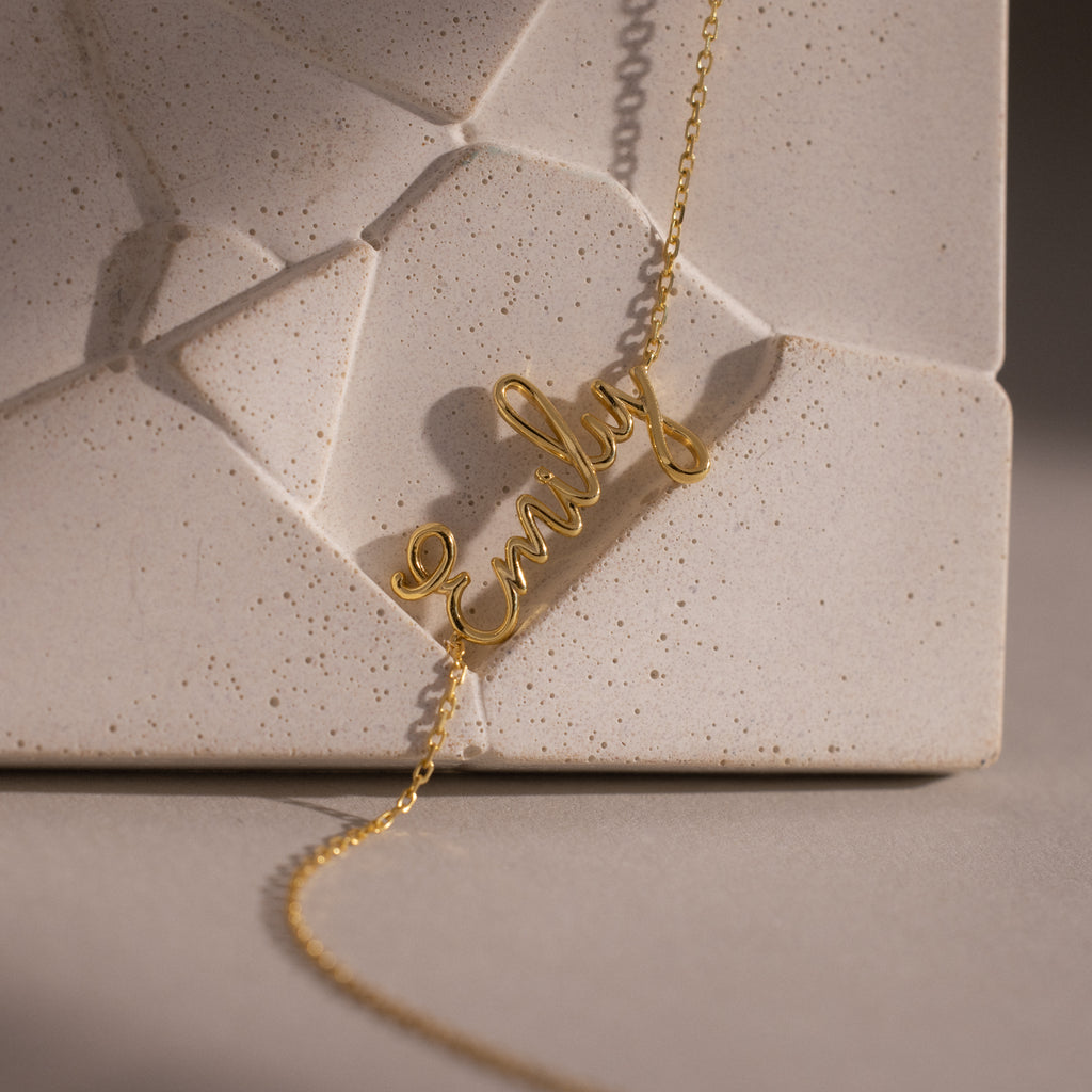 Cursive Name Bracelet in 18K Gold shown resting on a textured, geometric beige surface—a stunning custom jewelry piece that adds a personal touch to any look.