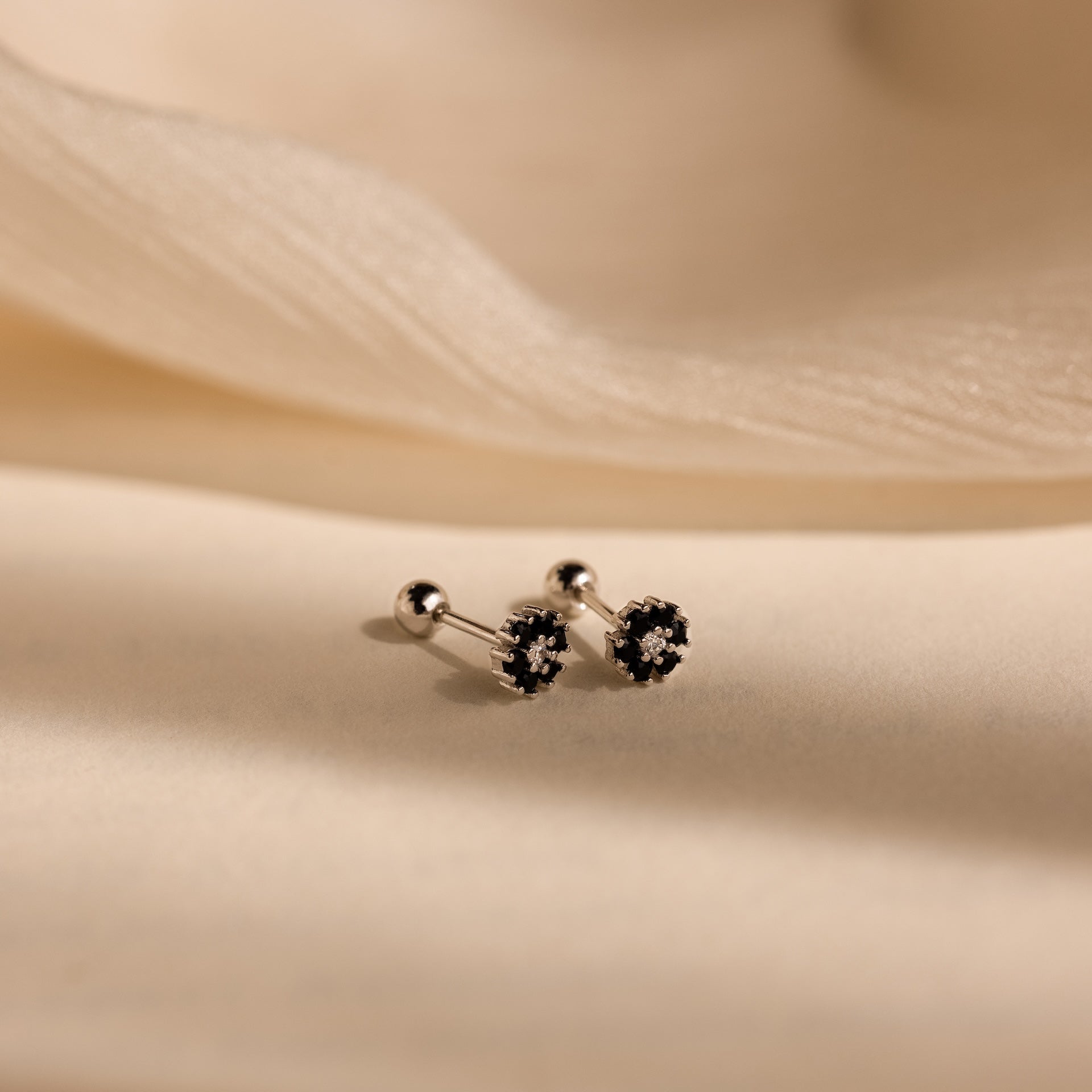 The Onyx Flower Studs in Silver, featuring dark, flower-shaped accents, are displayed elegantly on a soft beige fabric background.
