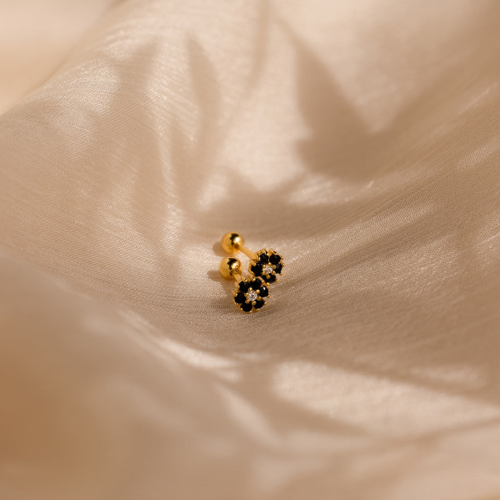 The Onyx Flower Studs, with gold and black onyx in a delicate flower shape, rest on beige satin—perfect dainty earrings for any occasion.
