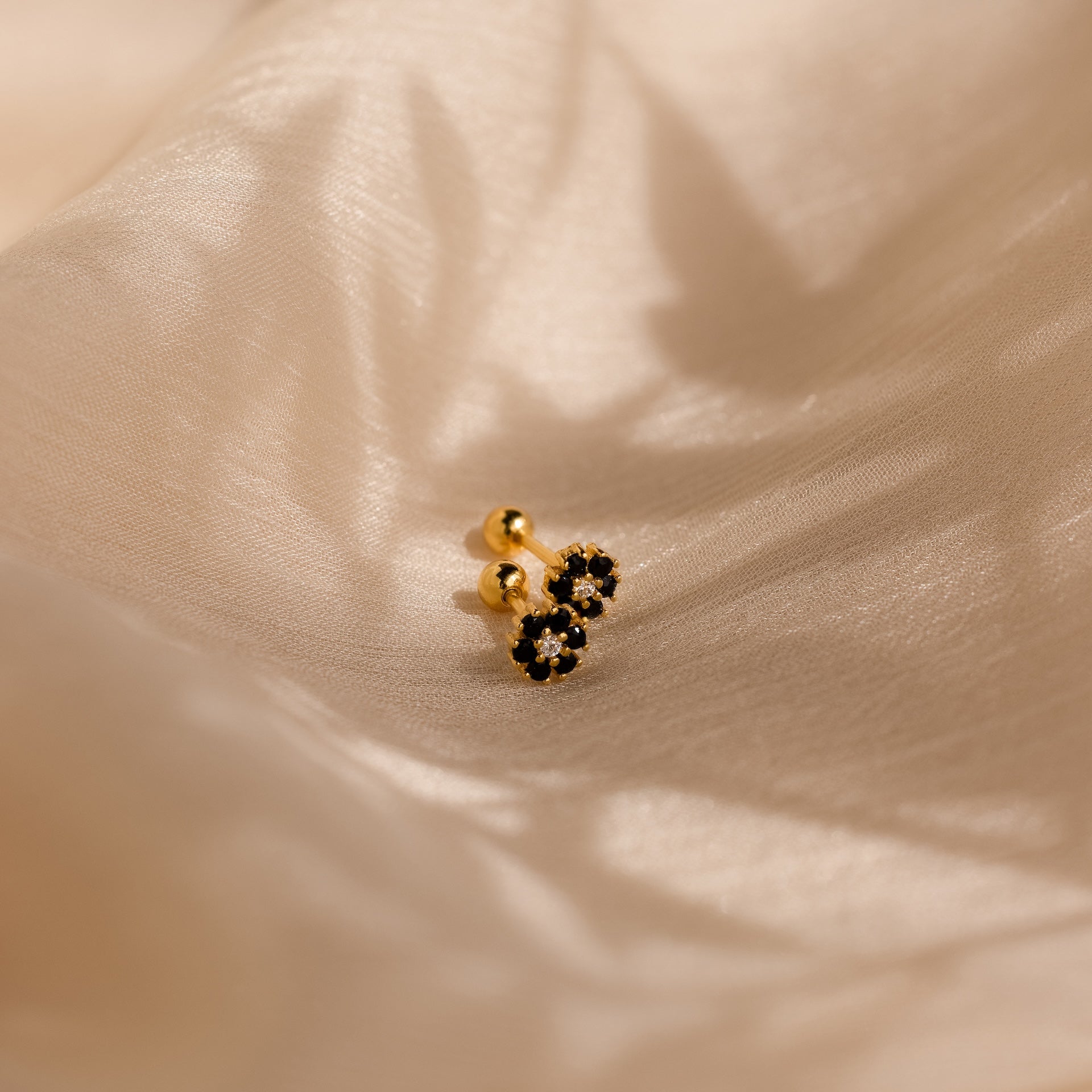 The Onyx Flower Studs, with gold and black onyx in a delicate flower shape, rest on beige satin—perfect dainty earrings for any occasion.