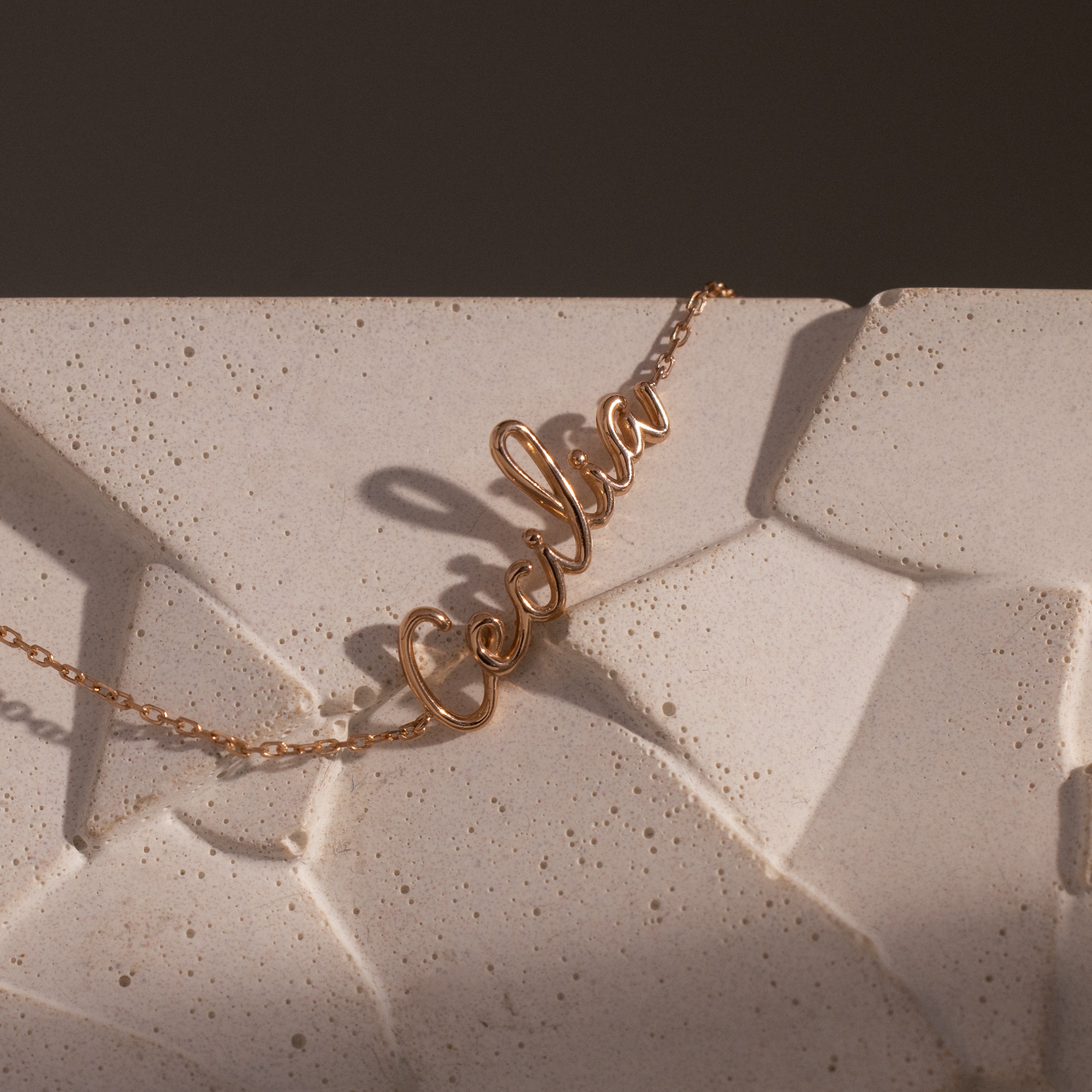 The Cursive Name Bracelet in Rose Gold, featuring “Cecilia” in elegant script, rests on a textured light beige stone surface.