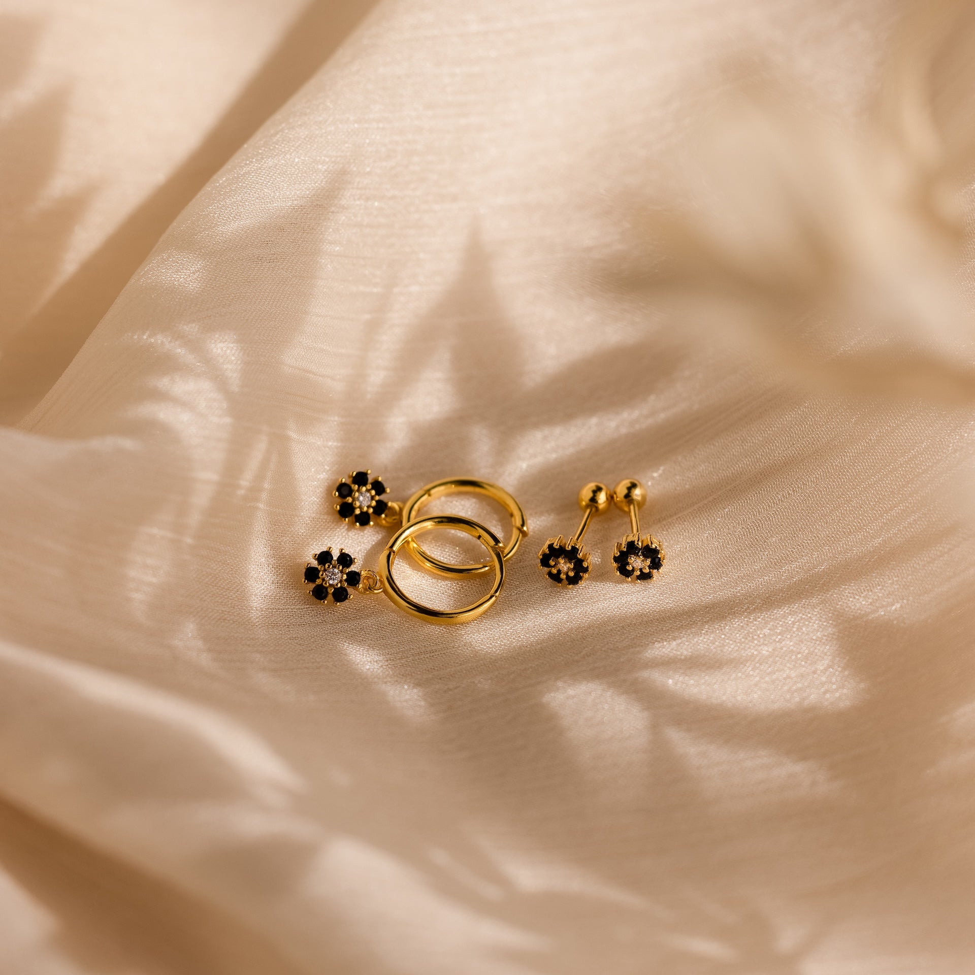Onyx Flower Studs and gold rings lie on beige silky fabric, illuminated by gentle light and soft shadows.