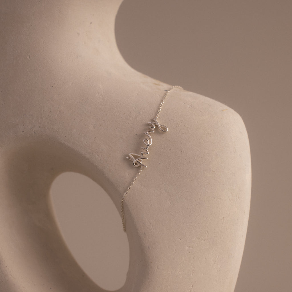 The Cursive Name Bracelet in Sterling Silver displays the word "merci" in elegant script, gracefully draped on a smooth, abstract sculpture featuring a round opening.