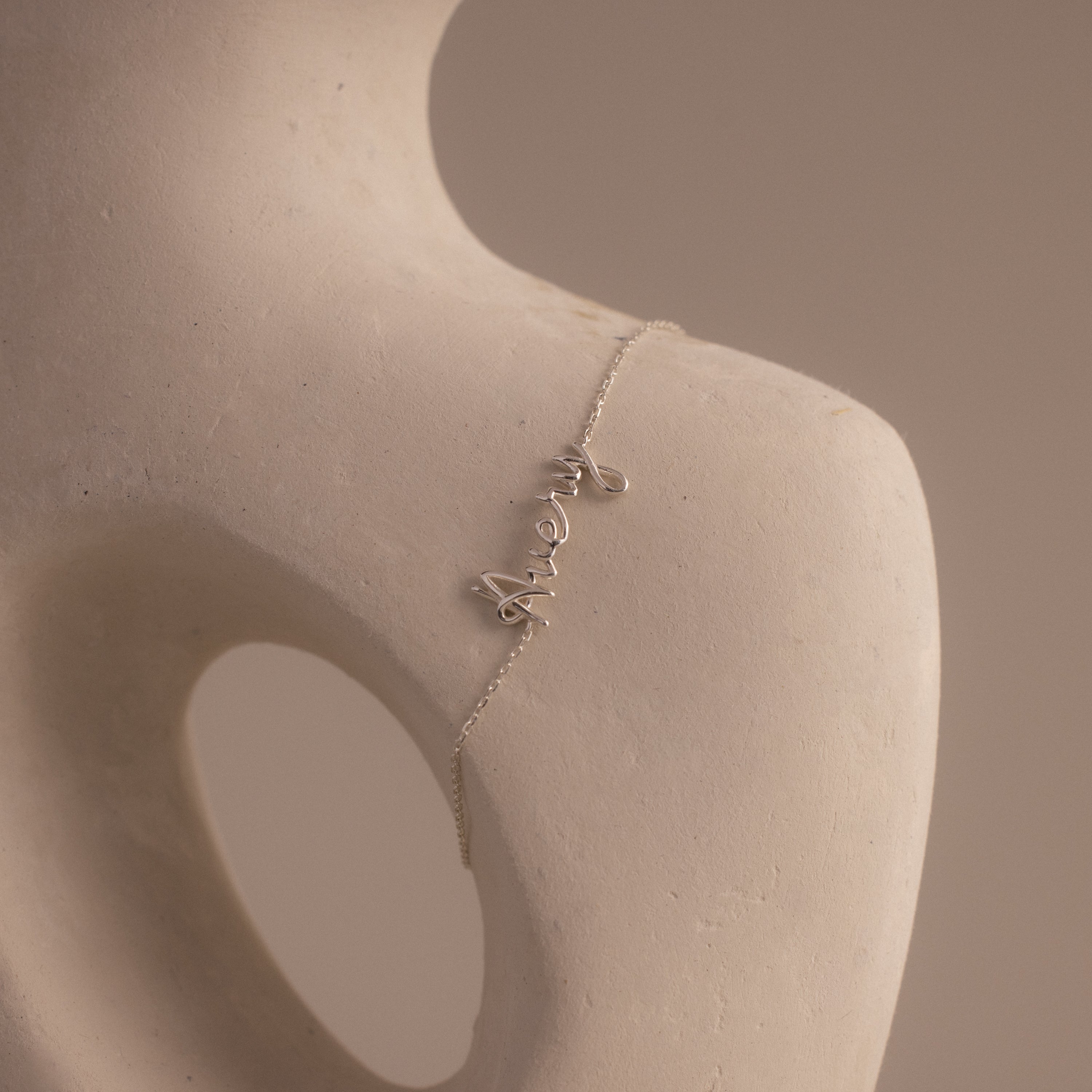 The Cursive Name Bracelet in Sterling Silver displays the word "merci" in elegant script, gracefully draped on a smooth, abstract sculpture featuring a round opening.