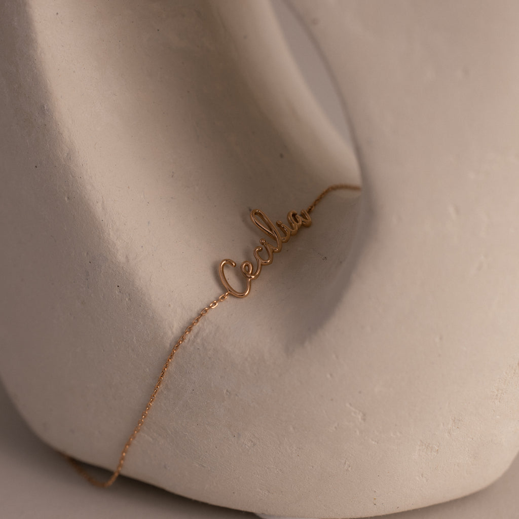 A rose gold cursive name bracelet with "Cecilia" elegantly displayed, draped over a smooth, abstract white sculpture.