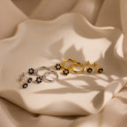 Three pairs of floral earrings, including Onyx Flower Studs in Silver and gold diamond center earrings, are displayed on a wavy-edged cream ceramic plate.