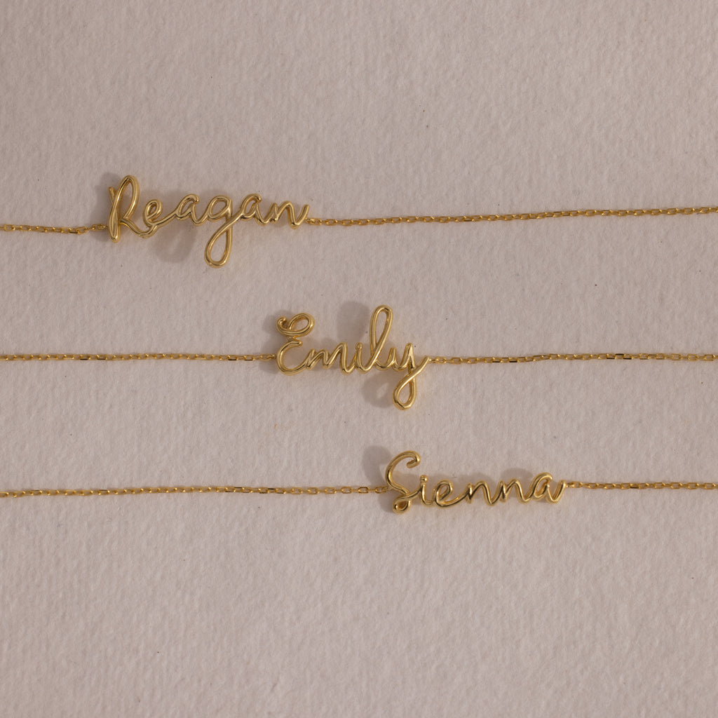 An 18K gold cursive name bracelet featuring custom names—Reagan, Emily, and Sienna—displayed on a beige background.