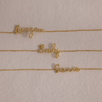 An 18K gold cursive name bracelet featuring custom names—Reagan, Emily, and Sienna—displayed on a beige background.