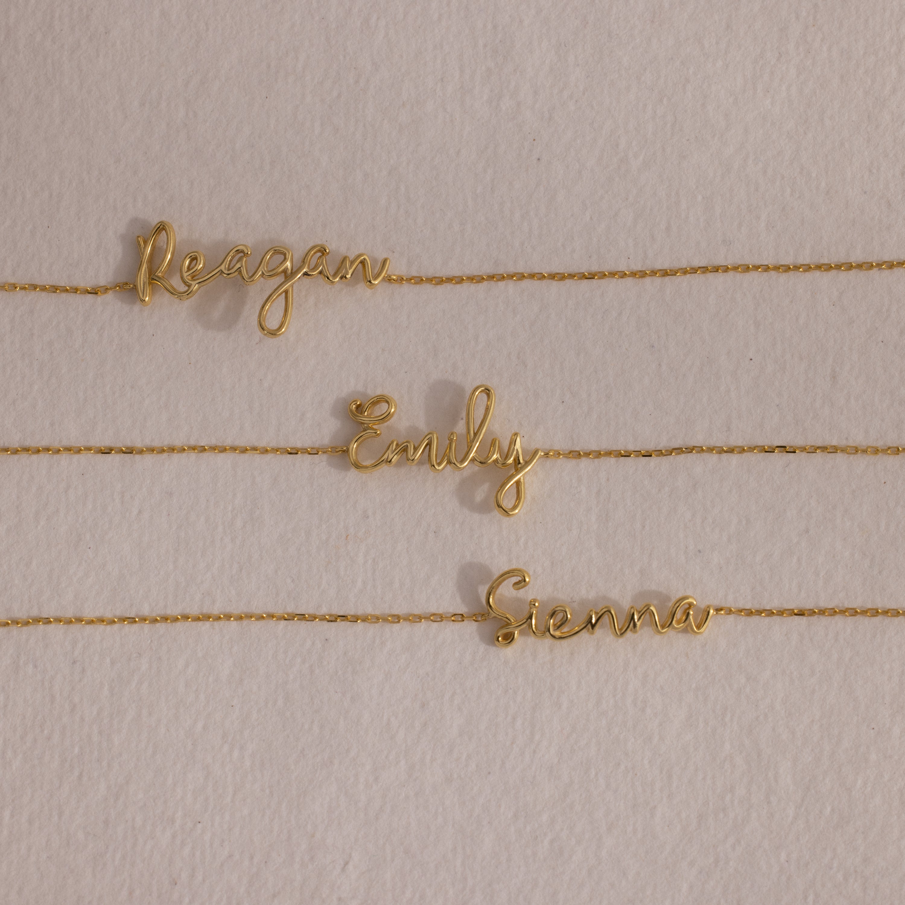 An 18K gold cursive name bracelet featuring custom names—Reagan, Emily, and Sienna—displayed on a beige background.