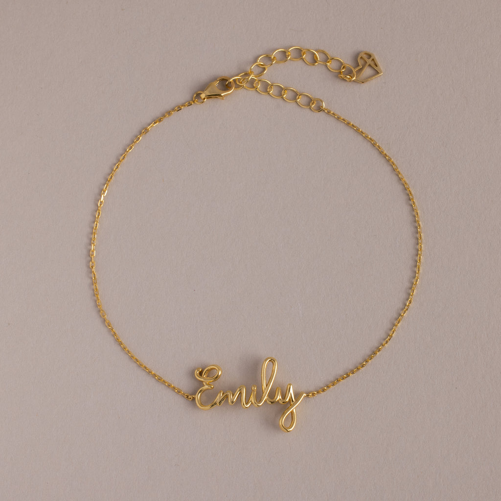 The Cursive Name Bracelet in 18K Gold features "Emily" in elegant script on a neutral background—a perfect personalized gift for custom name jewelry lovers.