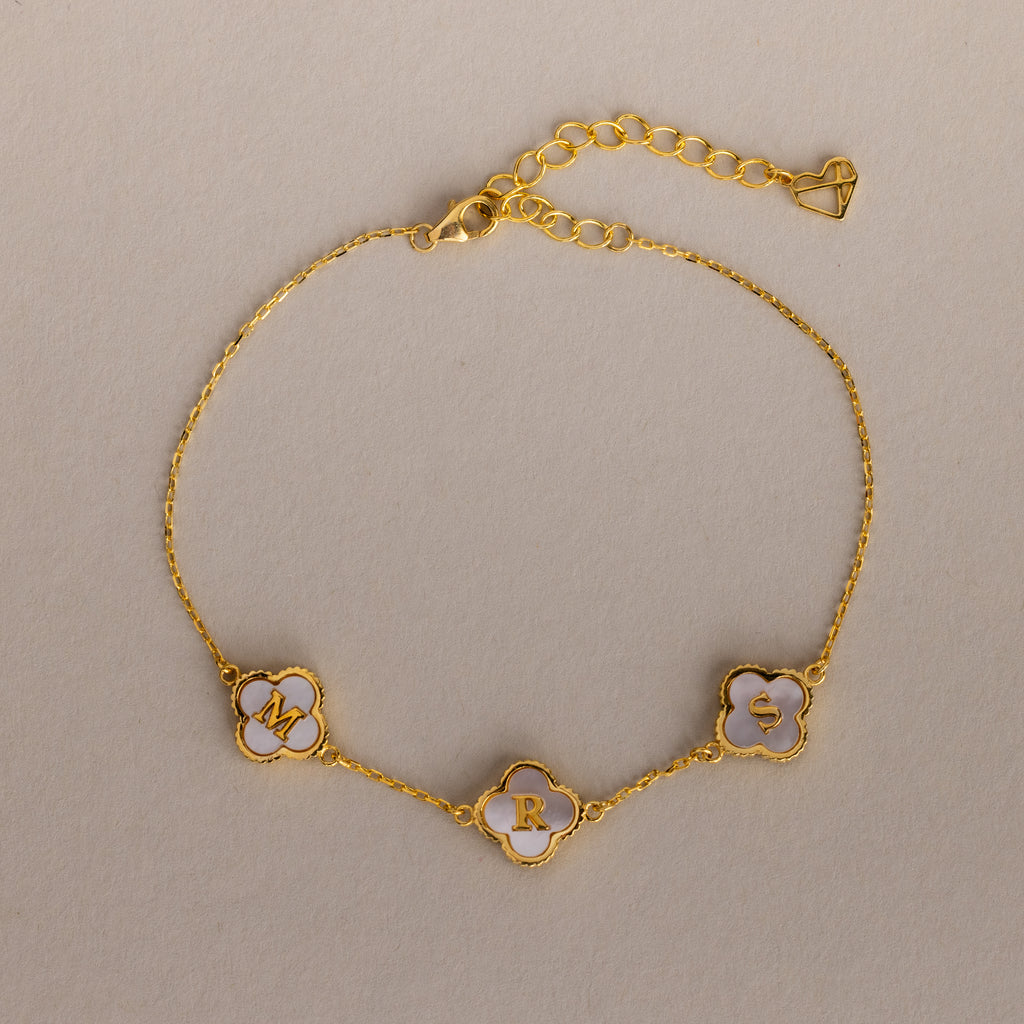 The Pearl Clover Letter Bracelet in 18K Gold features three white mother of pearl clover charms with the letters M, R, and S on a neutral background.