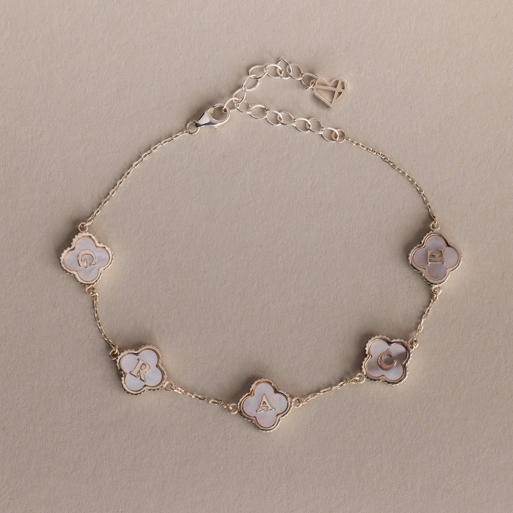 The Pearl Clover Letter Bracelet in Sterling Silver features five white mother of pearl clover-shaped charms, each with a letter from "GRACE," displayed on a beige background.