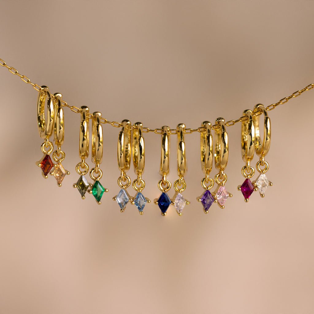 Nine Birthstone Drop Huggies featuring colorful gemstone charms on thin gold chains, perfect for personalized jewelry. Displayed against a neutral background.
