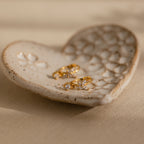 The Butterfly Drop Huggies in 18K Gold, featuring delicate butterfly designs, rest in a heart-shaped ceramic dish on a beige surface.