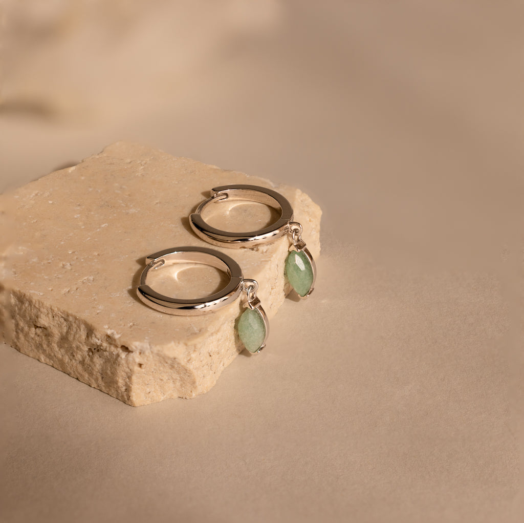 The Jade Marquise Huggies, featuring silver hoops with marquise jade charms, are displayed on a textured beige stone block for a unique botanical accent.