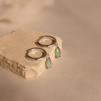 The Jade Marquise Huggies, featuring silver hoops with marquise jade charms, are displayed on a textured beige stone block for a unique botanical accent.