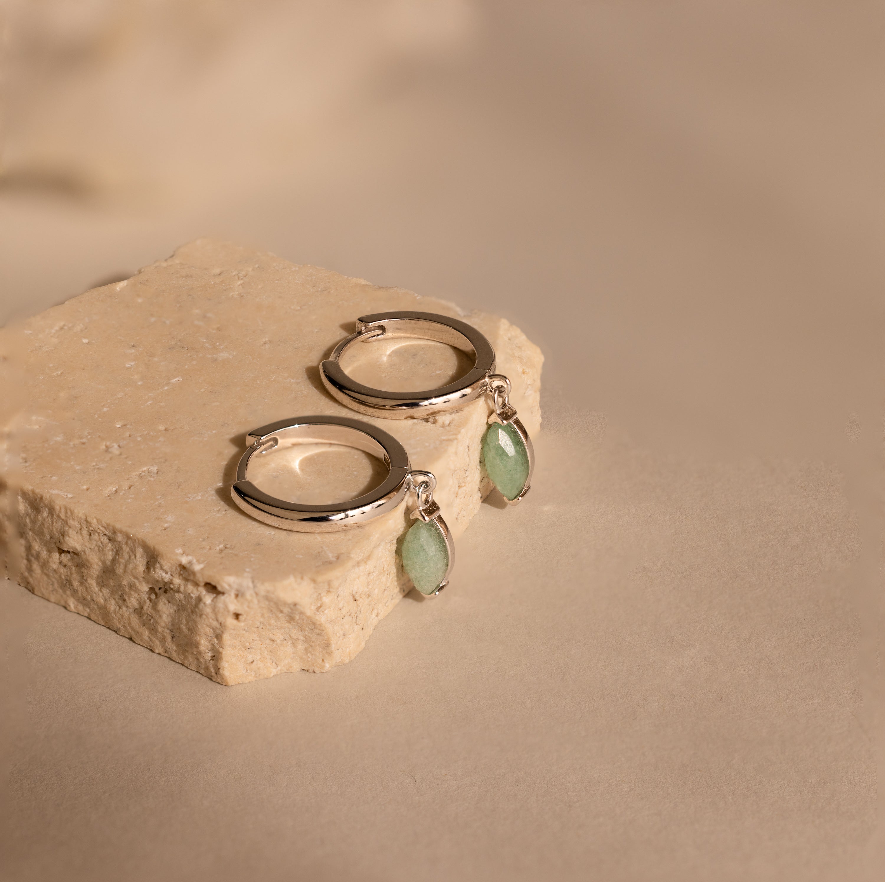 The Jade Marquise Huggies, featuring silver hoops with marquise jade charms, are displayed on a textured beige stone block for a unique botanical accent.