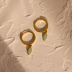 Jade Marquise Huggies—gold hoop earrings featuring oval green stones reminiscent of jade—displayed on a textured beige surface in warm lighting.