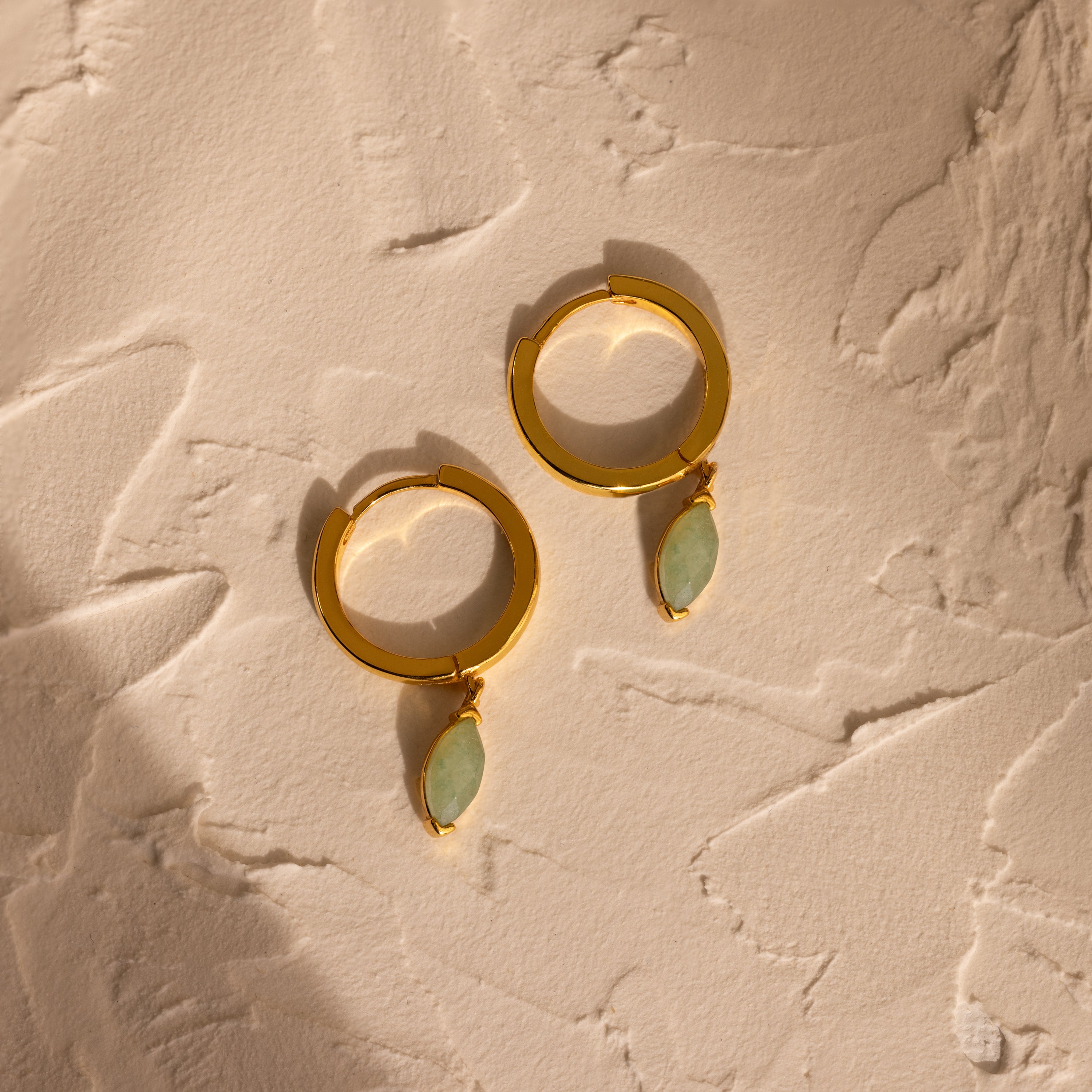 Jade Marquise Huggies—gold hoop earrings featuring oval green stones reminiscent of jade—displayed on a textured beige surface in warm lighting.