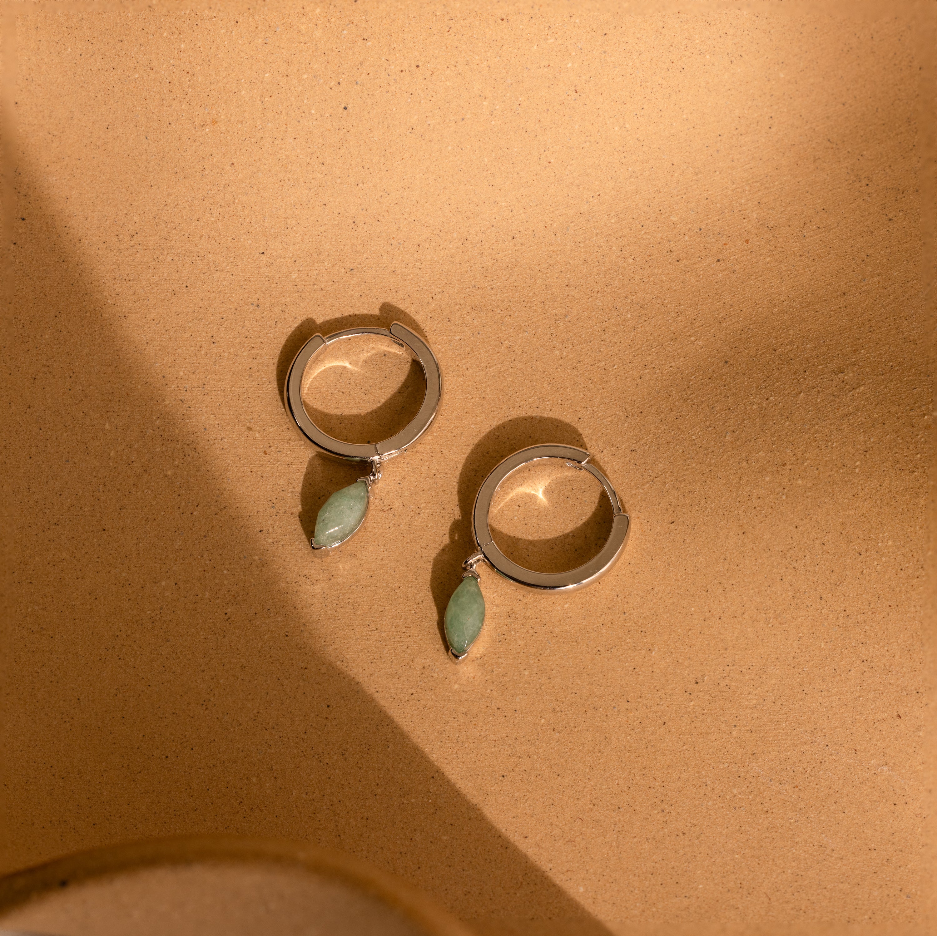 Jade Marquise Huggies in Silver displayed on a sandy beige background under soft light.