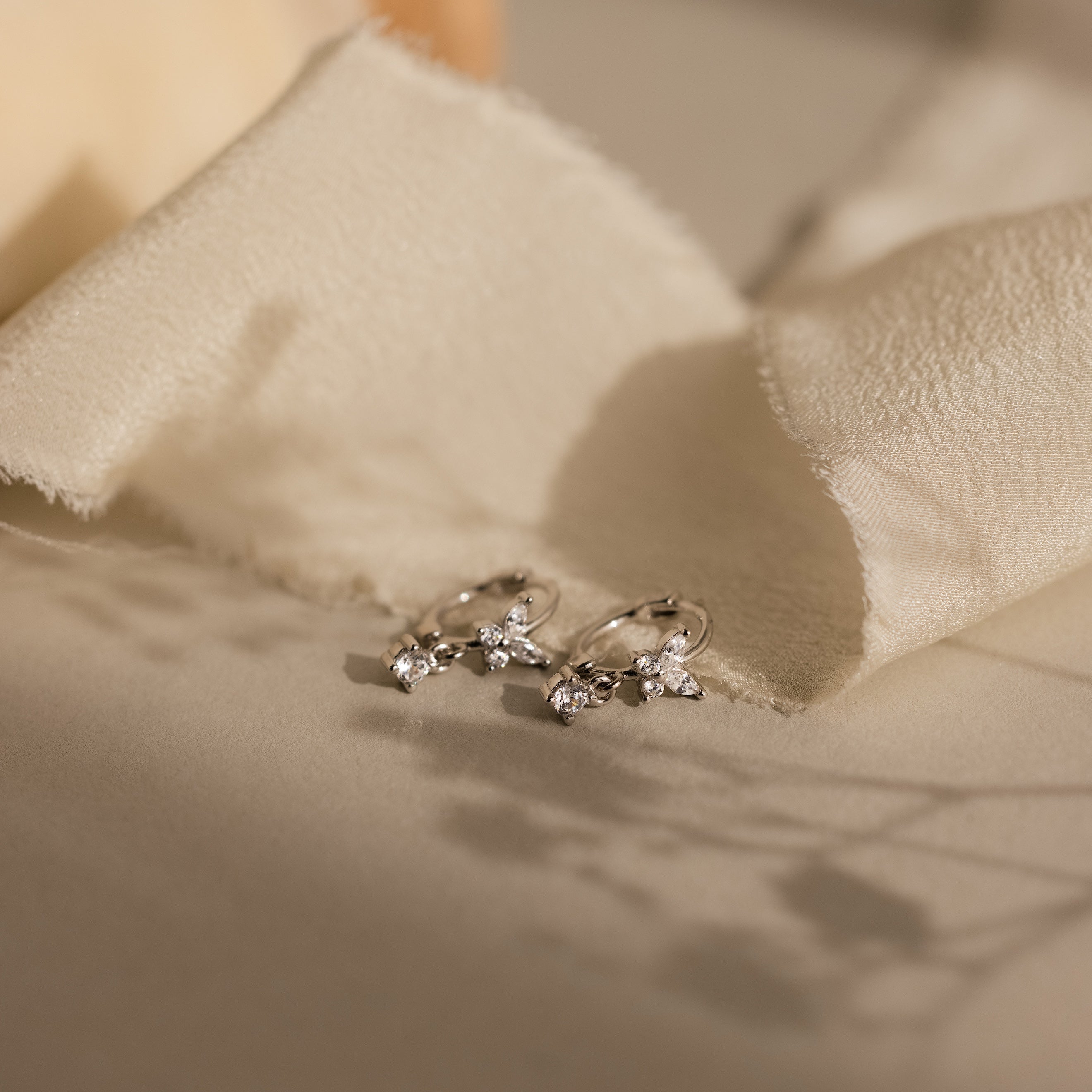 Butterfly Drop Huggies in Sterling Silver with small gemstones and diamond accents displayed on soft beige fabric with gentle shadows.