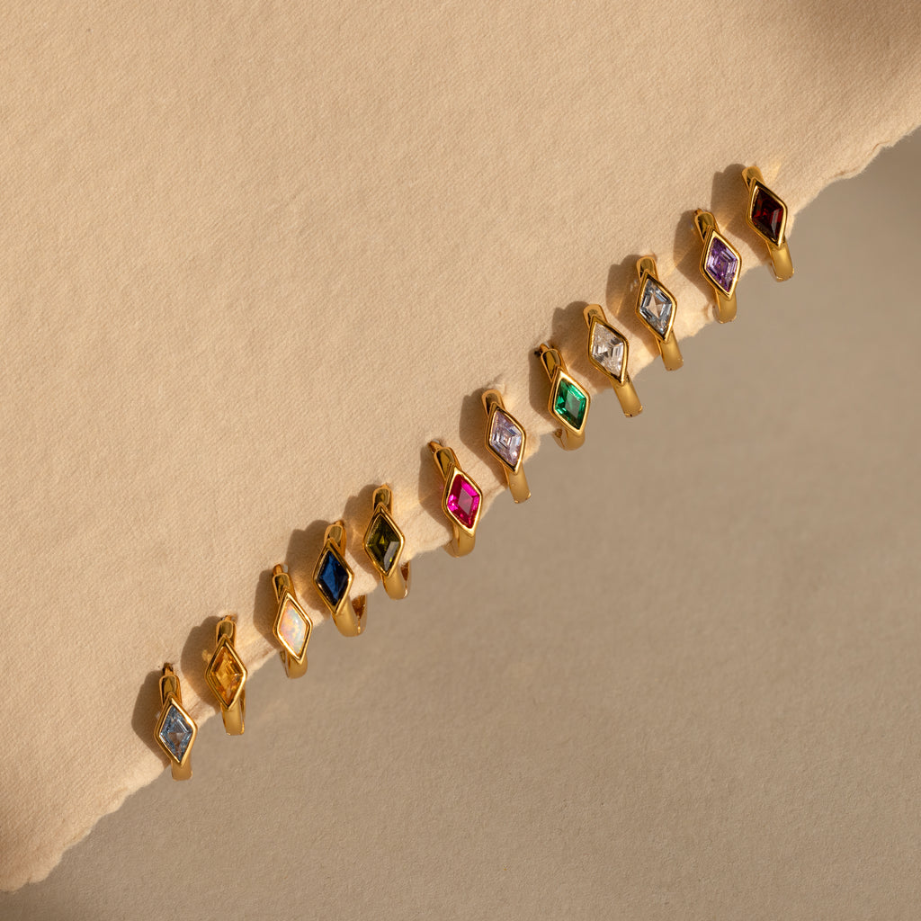 A full lineup of gold huggie earrings each set with a different colored rhombus-shaped birthstone, arranged diagonally on a beige background showcasing all 12 birthstones from garnet to zircon.