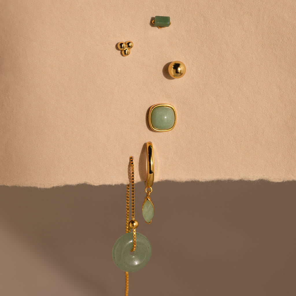 Jade Marquise Huggies in 18K Gold, along with green gemstone jewelry, arranged vertically on a textured beige surface.