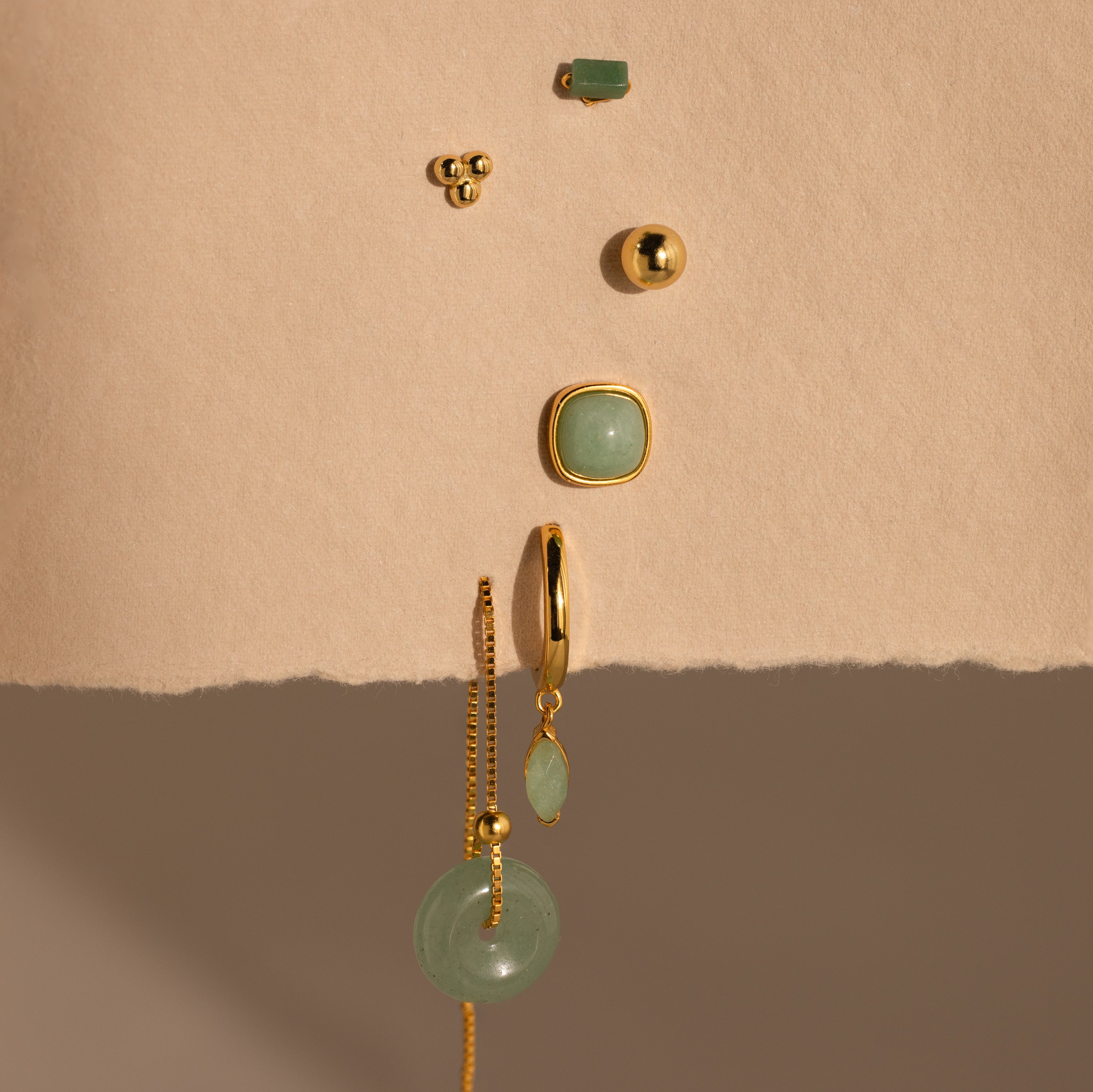 Jade Marquise Huggies in 18K Gold, along with green gemstone jewelry, arranged vertically on a textured beige surface.