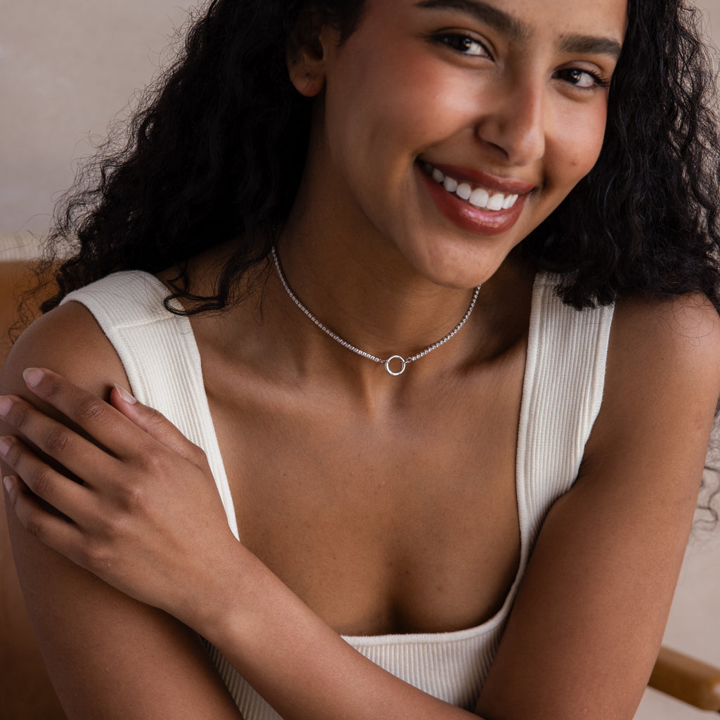 A smiling woman with curly hair wears a white top and the Diamond Tennis Clasp Necklace in White Gold, featuring a rolo clasp, while seated with her arms crossed.