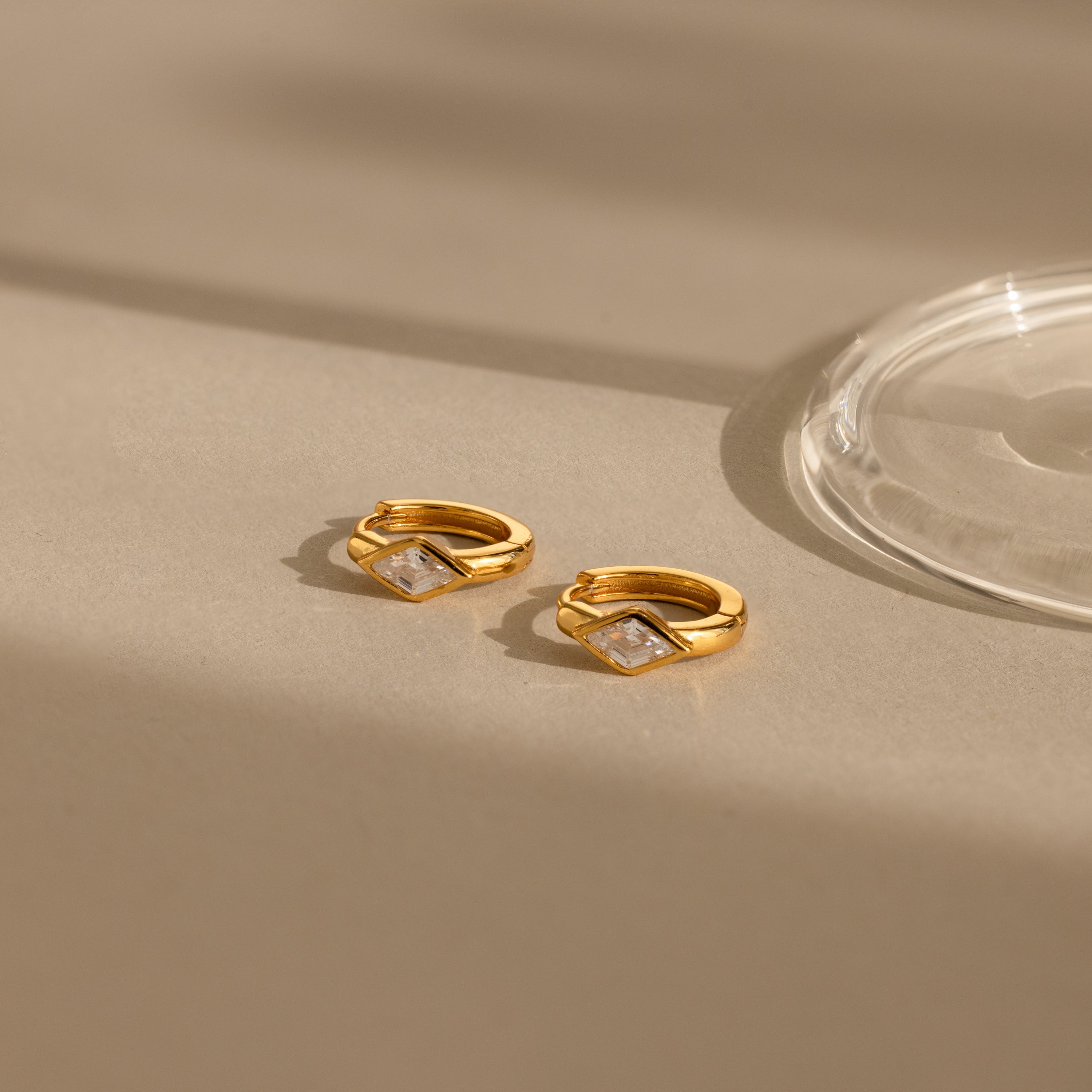Rhombus Diamond Huggies—two gold huggie earrings with square clear stones, shown on a beige surface by a glass object. These customizable earrings add subtle elegance to any look.