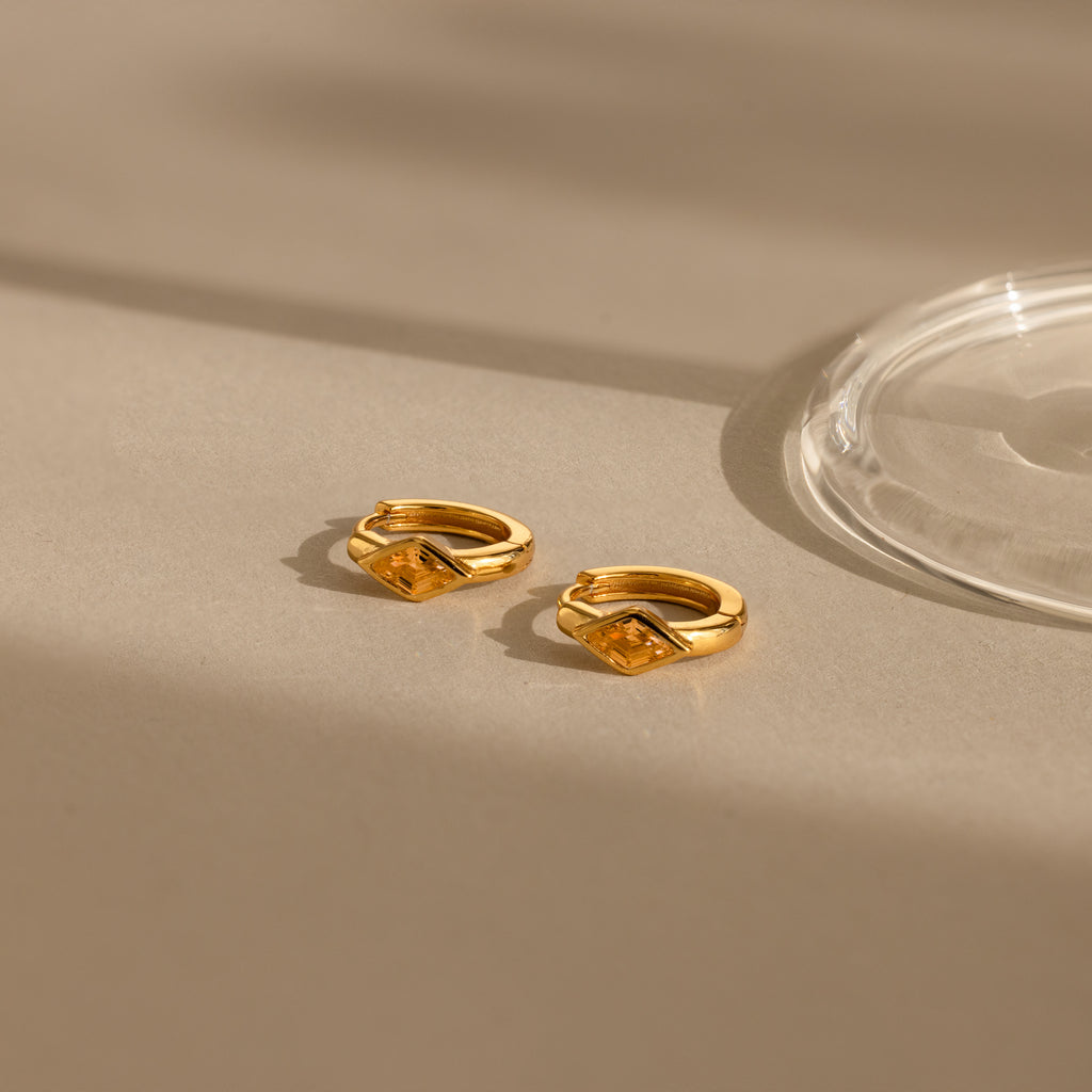 The Rhombus Topaz Huggies, gold hoop earrings with geometric design, are displayed on a neutral beige surface in soft light and shadows.