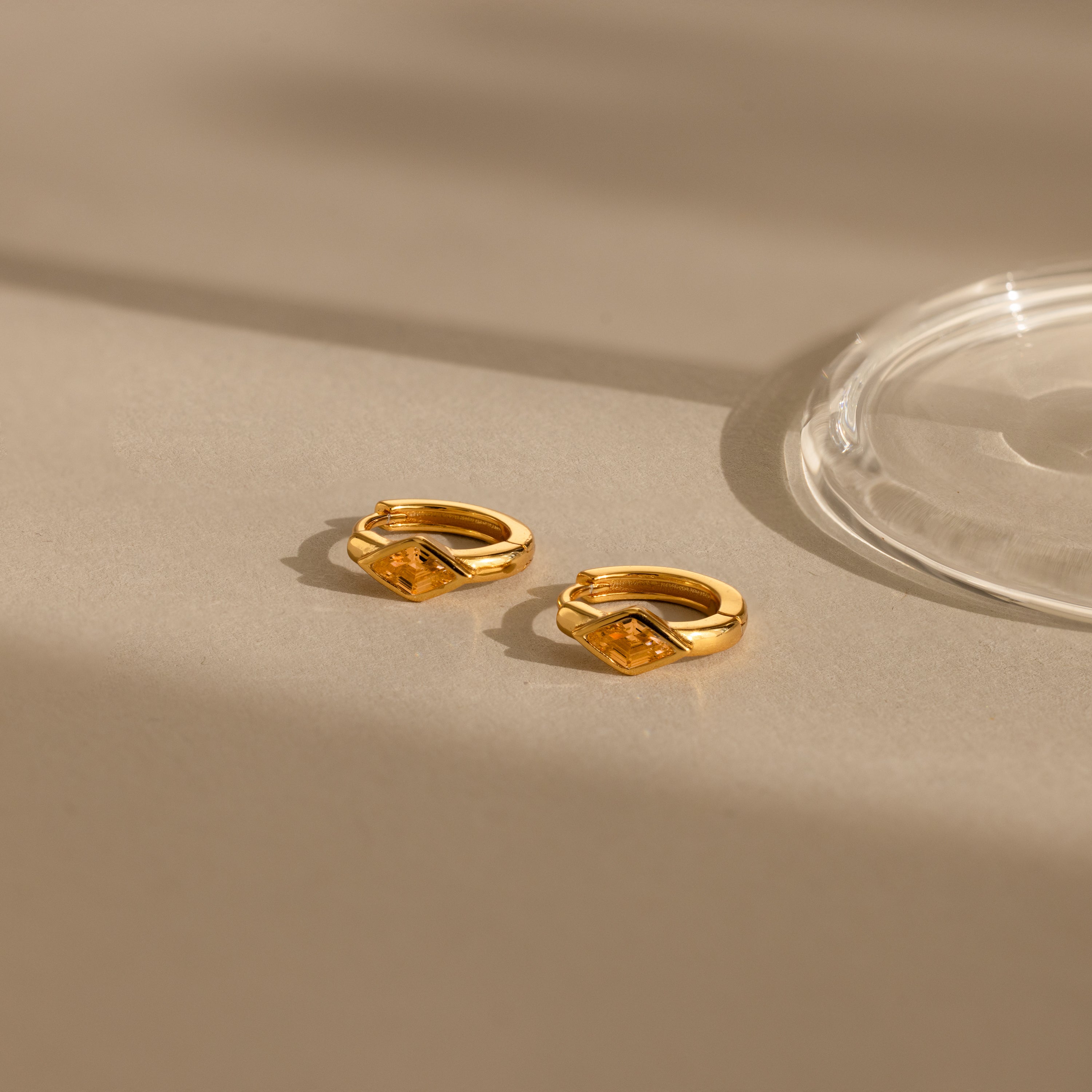 The Rhombus Topaz Huggies, gold hoop earrings with geometric design, are displayed on a neutral beige surface in soft light and shadows.