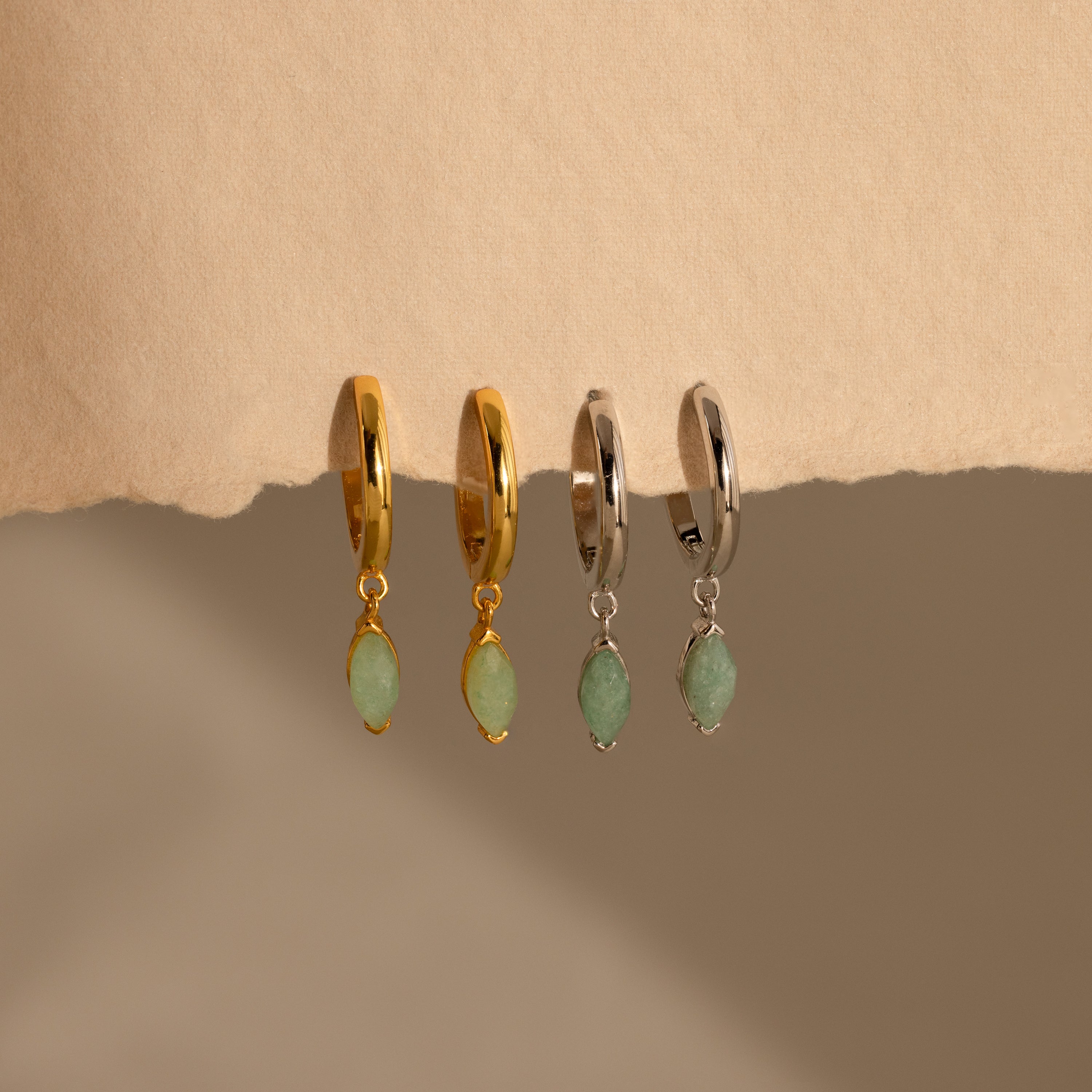 Two pairs of Jade Marquise Huggies in gold and silver, featuring green marquise jade stones, are displayed on a beige textured surface.