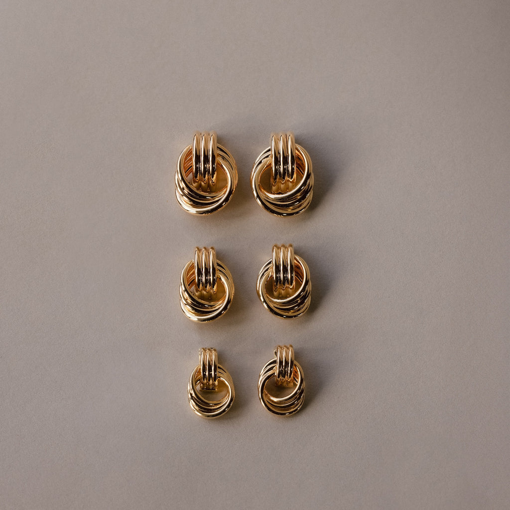 Six pairs of Micro Triple Knot Earrings, featuring intertwined gold rings, are arranged in two neat columns on a plain beige background—a subtle nod to quiet luxury.
