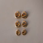 Six pairs of Micro Triple Knot Earrings, featuring intertwined gold rings, are arranged in two neat columns on a plain beige background—a subtle nod to quiet luxury.