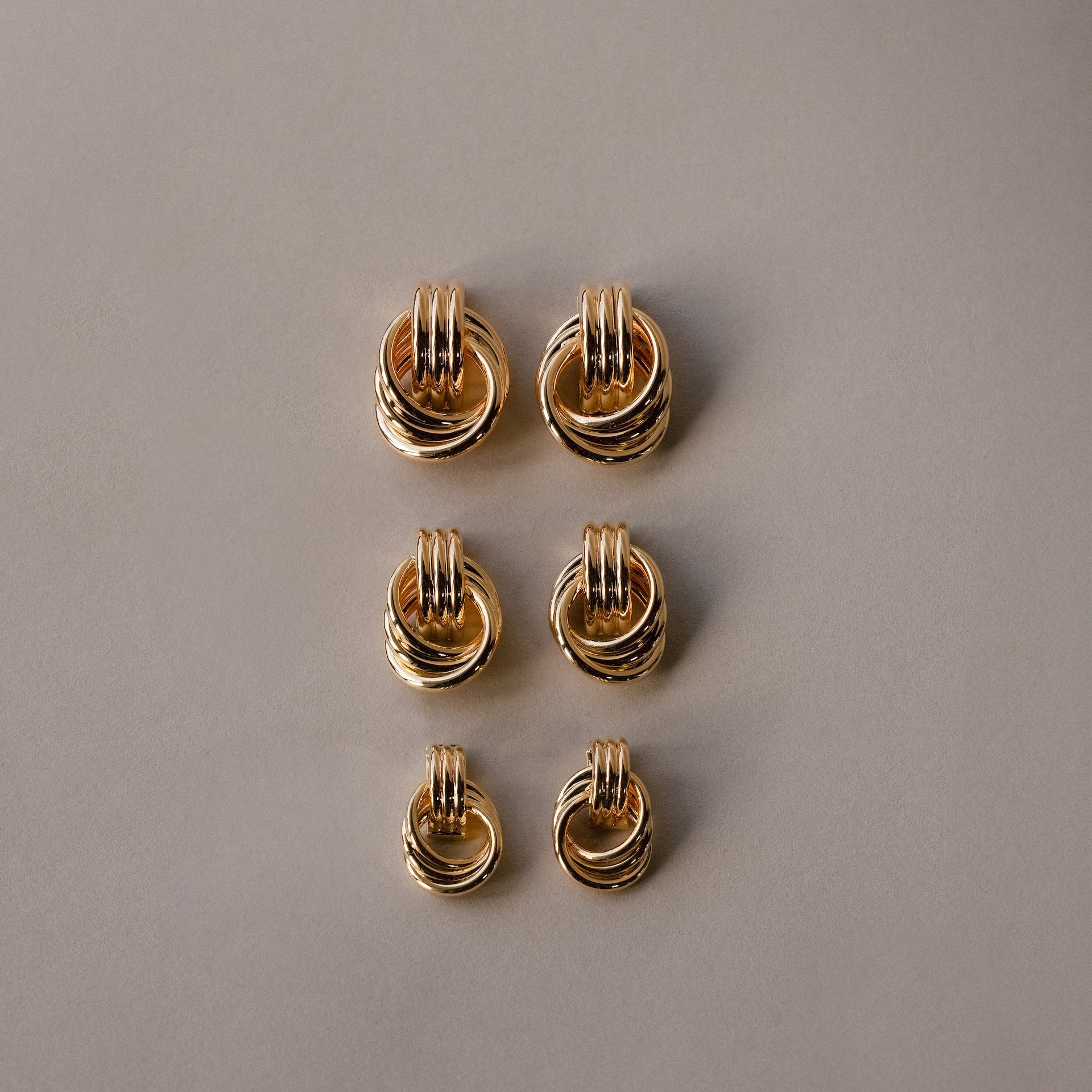 Six pairs of Micro Triple Knot Earrings, featuring intertwined gold rings, are arranged in two neat columns on a plain beige background—a subtle nod to quiet luxury.