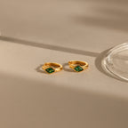 Rhombus Emerald Huggies—customizable gold hoop earrings with green rhombus gemstones—rest on a beige surface, softly illuminated by natural light.
