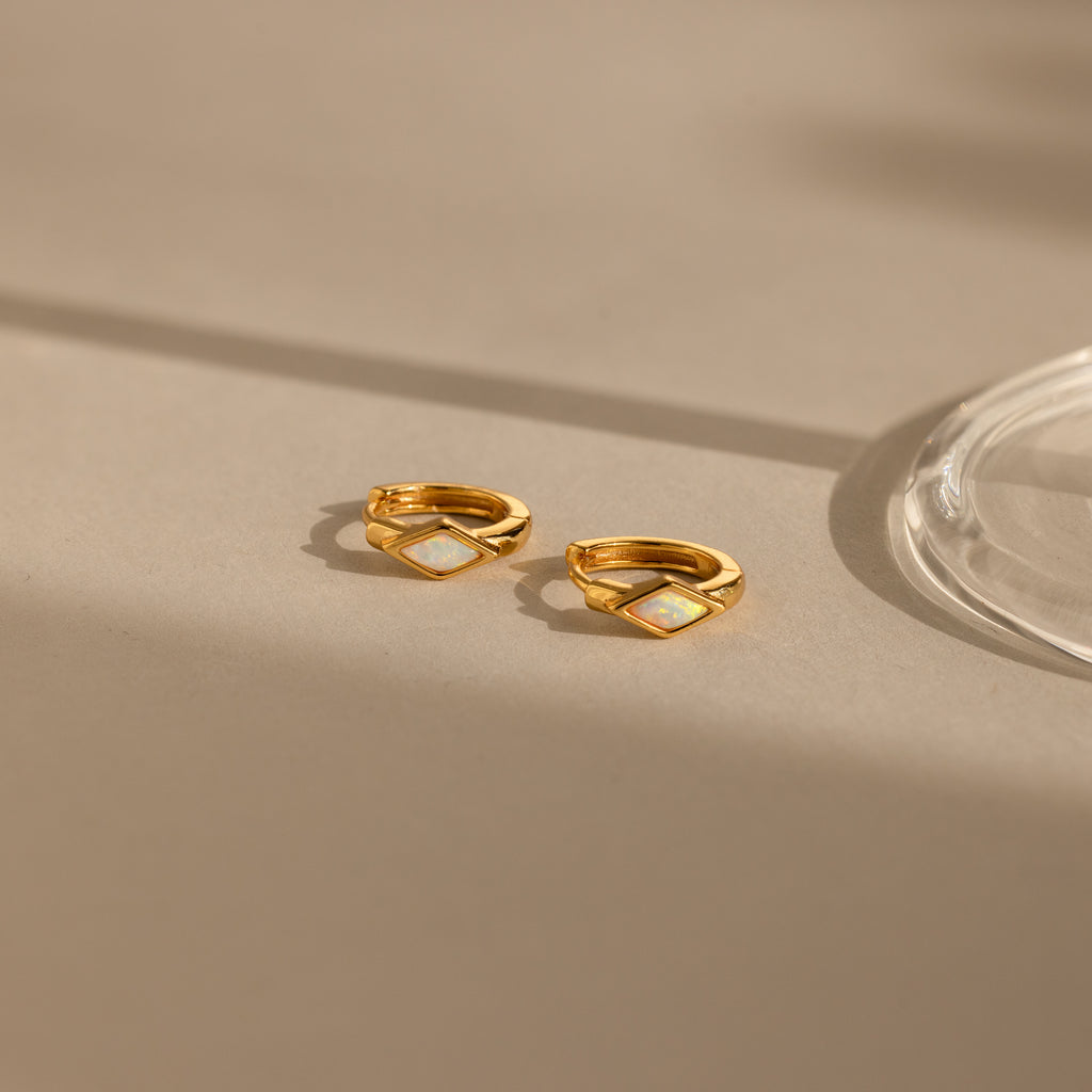 The Rhombus Opal Huggies are gold hoop earrings featuring diamond-shaped white opal inlays, offering a modern twist on classic gemstone hoops, elegantly presented on a beige surface with gentle shadows.