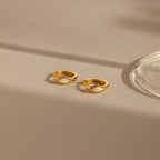 The Rhombus Opal Huggies are gold hoop earrings featuring diamond-shaped white opal inlays, offering a modern twist on classic gemstone hoops, elegantly presented on a beige surface with gentle shadows.