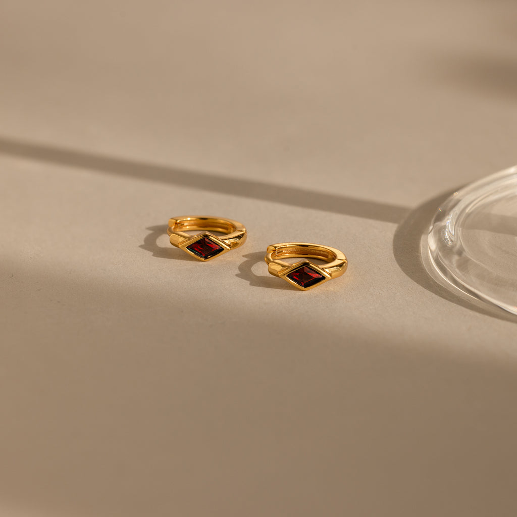 A close-up image of a pair of gold huggie earrings with rhombus-shaped deep red garnet stones, placed on a beige surface with soft natural shadows and a glass base on the right.