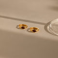 A close-up image of a pair of gold huggie earrings with rhombus-shaped deep red garnet stones, placed on a beige surface with soft natural shadows and a glass base on the right.