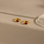 A close-up image of a pair of gold huggie earrings with rhombus-shaped deep red garnet stones, placed on a beige surface with soft natural shadows and a glass base on the right.