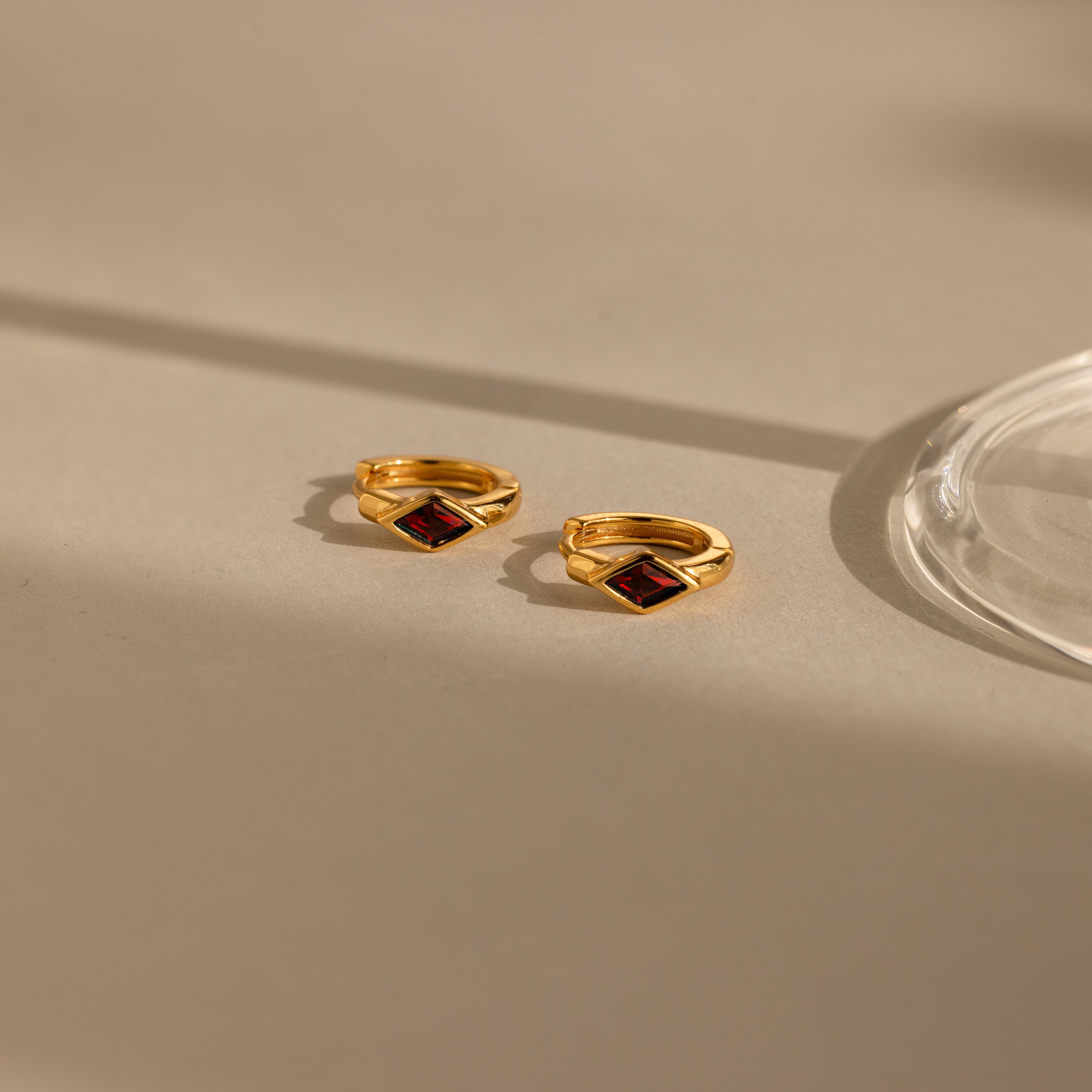 A close-up image of a pair of gold huggie earrings with rhombus-shaped deep red garnet stones, placed on a beige surface with soft natural shadows and a glass base on the right.