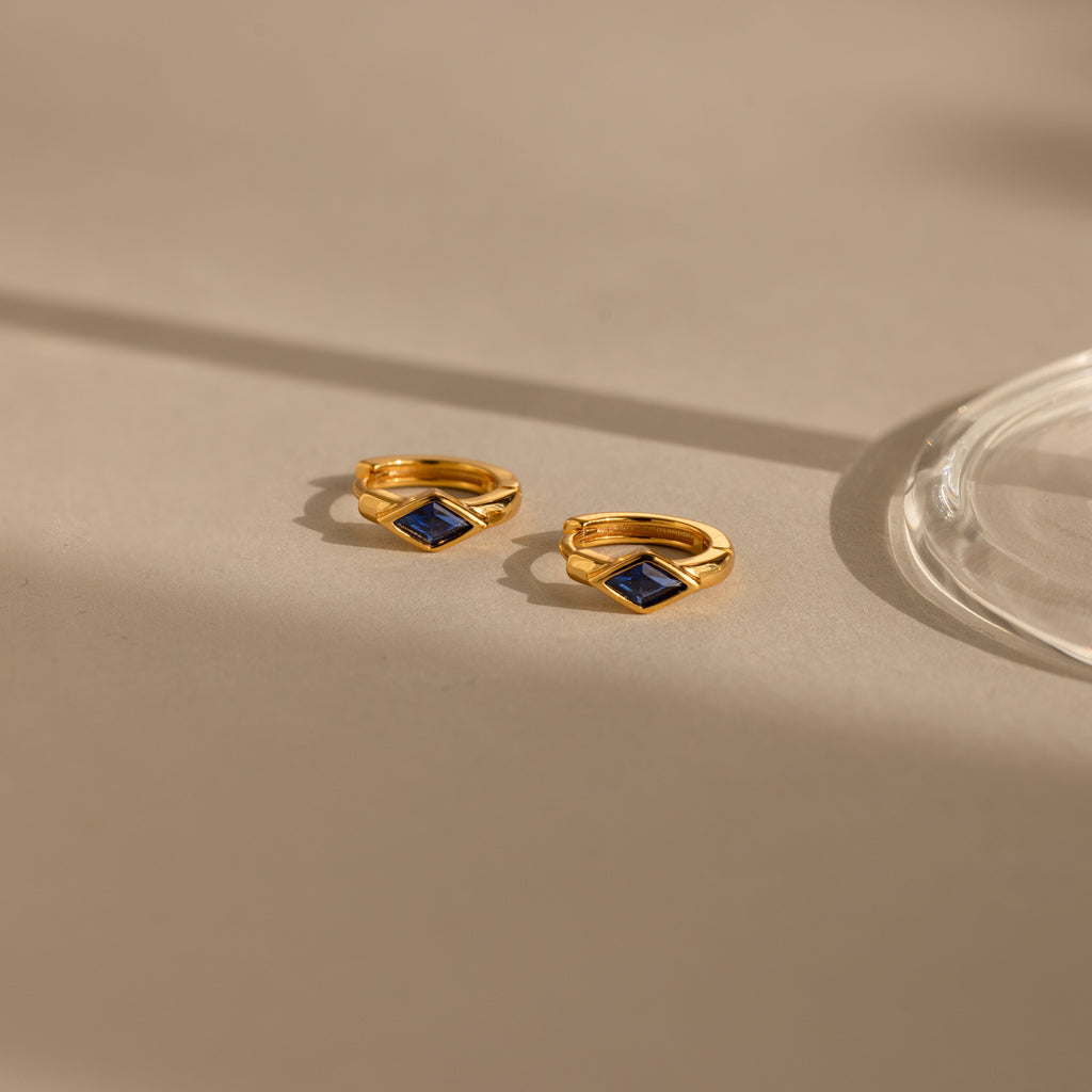 Two gold Rhombus Sapphire Huggies with square blue stones are displayed on a beige surface beside a glass object, highlighting the elegance of these gemstone huggie hoops.