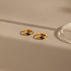 Two gold Rhombus Sapphire Huggies with square blue stones are displayed on a beige surface beside a glass object, highlighting the elegance of these gemstone huggie hoops.