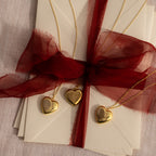 Flatlay image of our our Fingerprint Heart Locket Necklaces in Gold, laid on a stack of envelopes, tied to a red ribbon