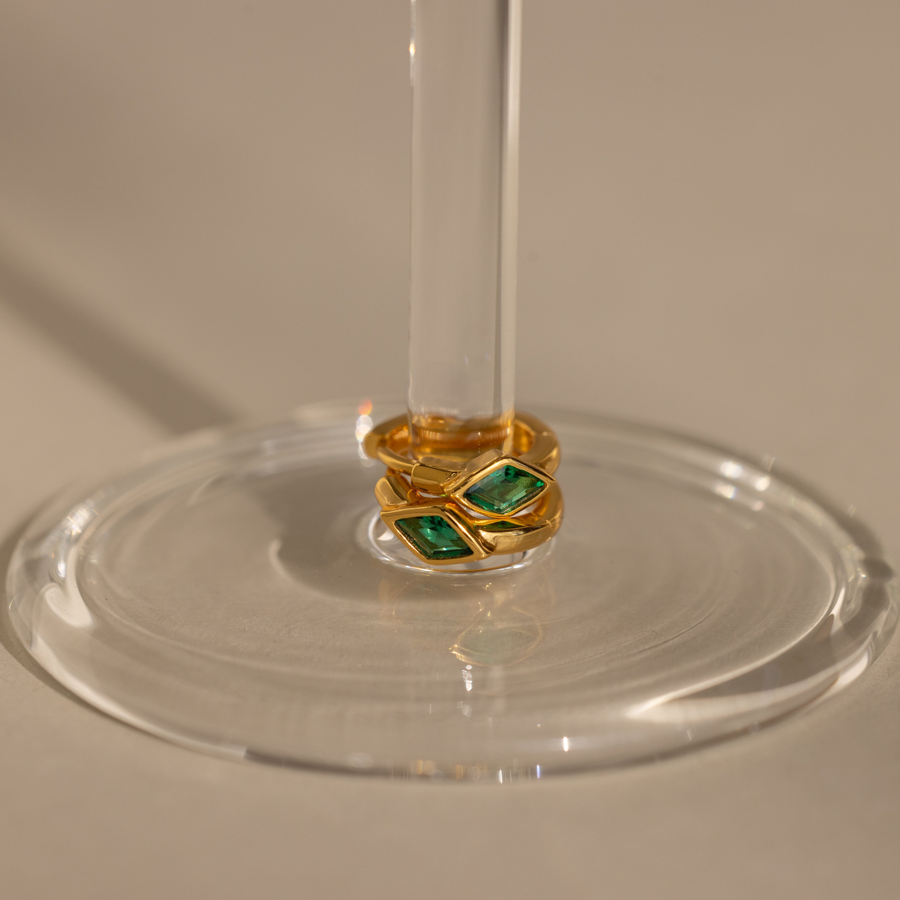 Two Rhombus Emerald Huggies with green stones are stacked around the stem of a clear wine glass placed on a beige surface.