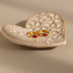 Two gold rings, inspired by Rhombus Ruby Huggies and set with red square gemstones, rest on a textured, heart-shaped ceramic dish.
