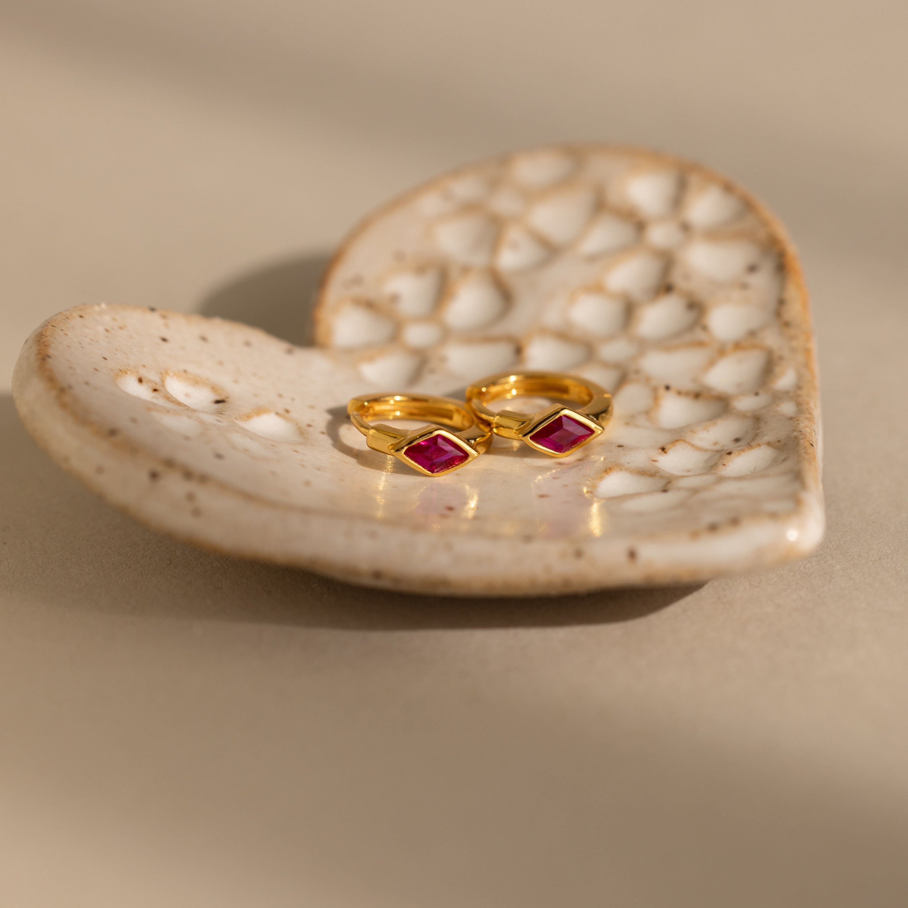 Two gold rings, inspired by Rhombus Ruby Huggies and set with red square gemstones, rest on a textured, heart-shaped ceramic dish.
