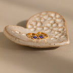 A pair of gold huggie earrings with rhombus-cut purple amethyst gemstones resting on a textured ceramic dish.
